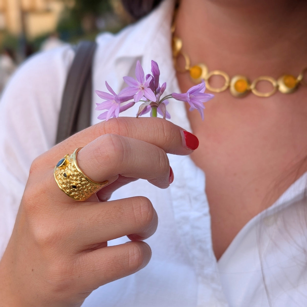 Anillo ancho dorado Neptuno de AMANAI con piedra azul, sujetando flores lilas al atardecer