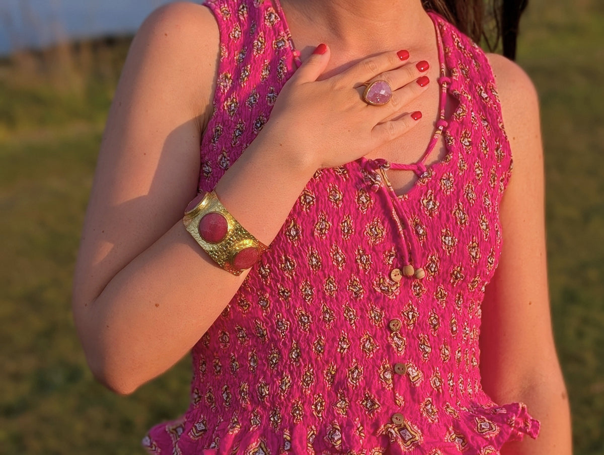 Detalle de anillo dorado con piedra natural rosa y brazalete dorado AMANAI sobre vestido fucsia