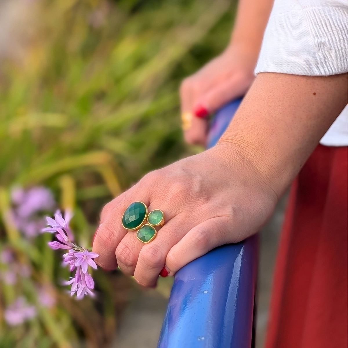 Detalle de anillo dorado con tres piedras verdes talladas de AMANAI, sujetando flores lilas