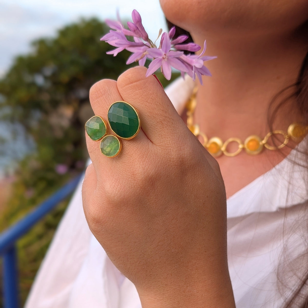 Anillo dorado con tres piedras naturales verdes de AMANAI sobre camisa blanca, con flores lilas