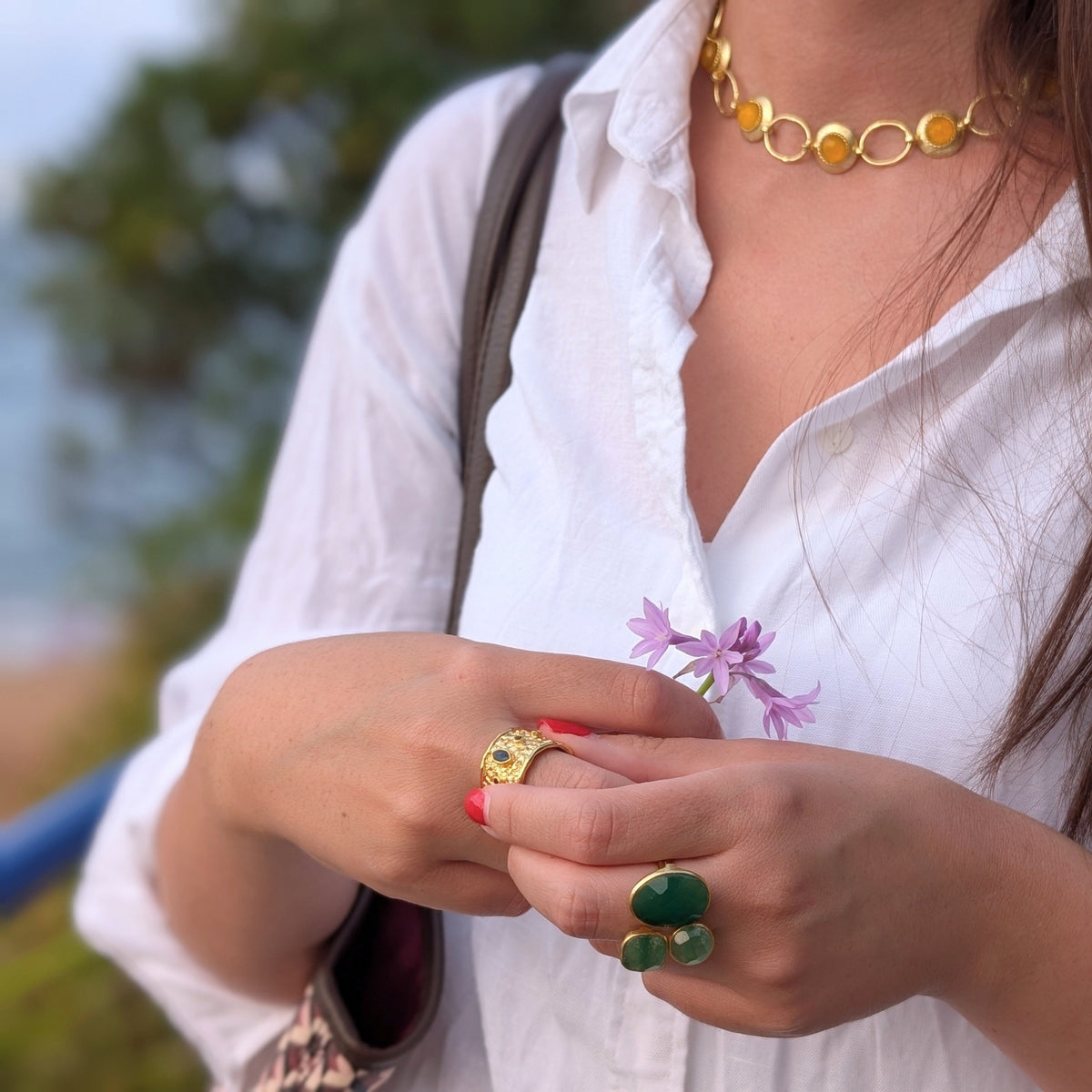 Manos con anillo ancho Neptuno y anillo de piedras verdes de AMANAI, con collar dorado de eslabones ámbar