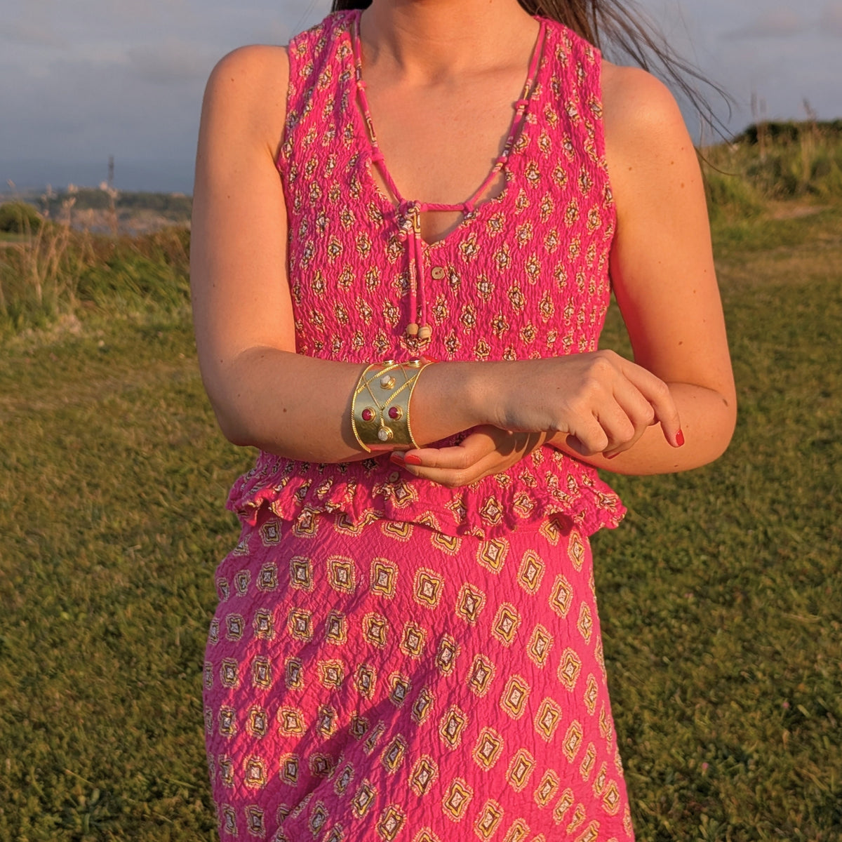 Mujer con brazalete ancho dorado Aurora de AMANAI con piedras naturales rojas, sobre vestido rosa al atardecer