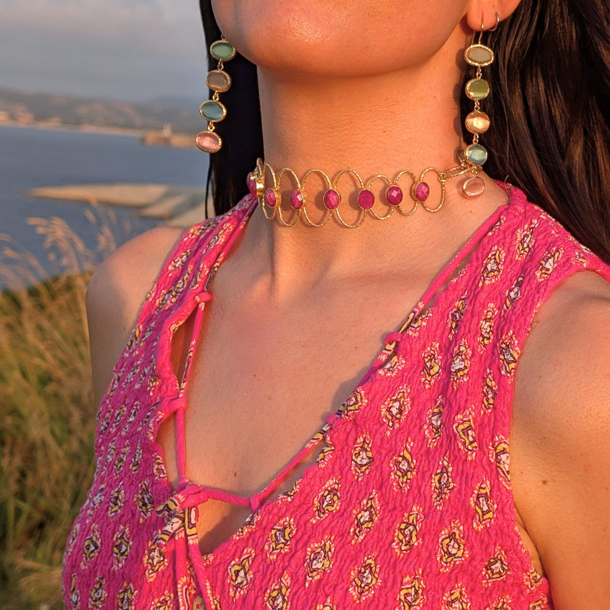 Mujer con choker dorado Coral de piedras fucsia y pendientes largos Prisma de AMANAI frente a la costa de Cantabria