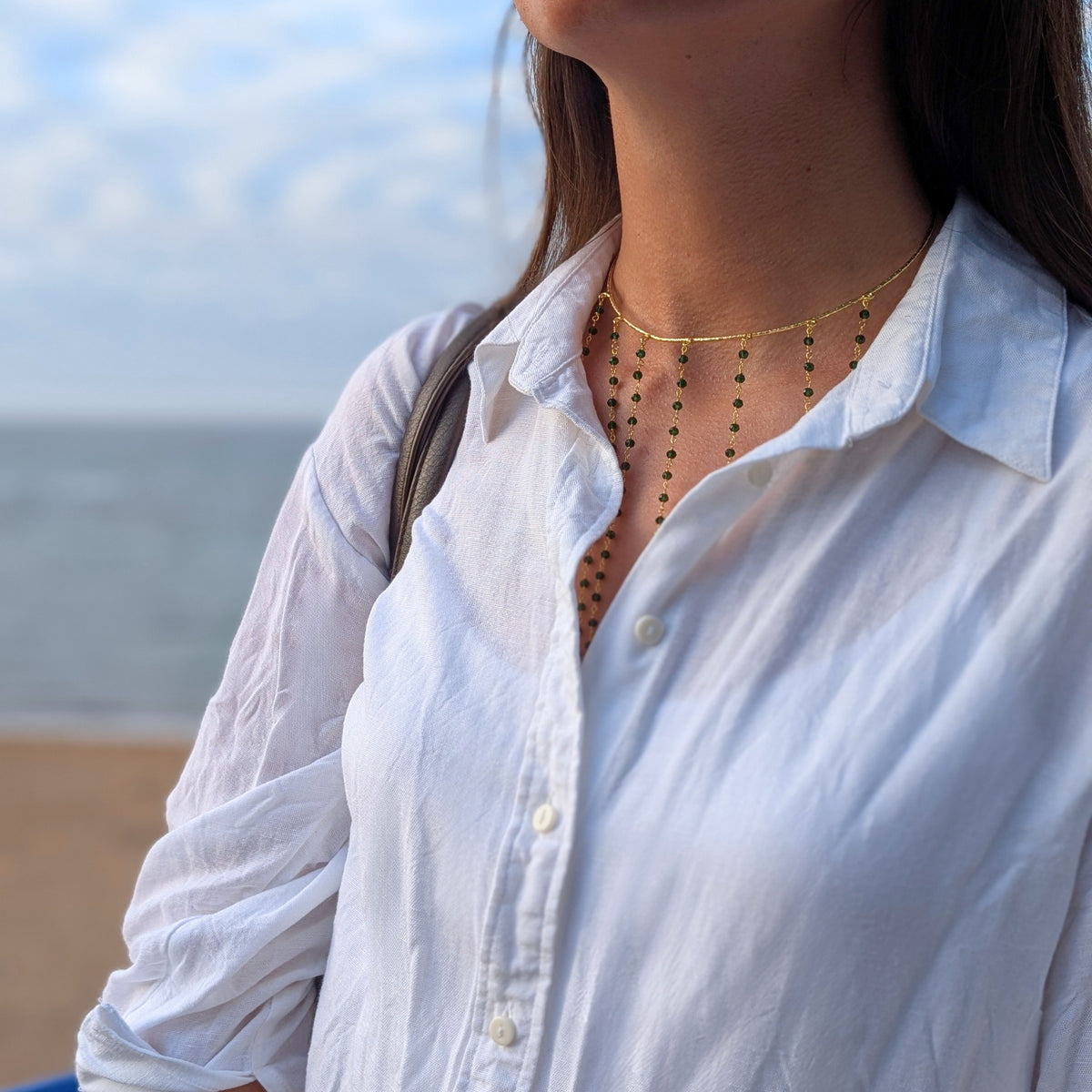 Mujer con choker dorado Isla de flecos con piedras verdes de AMANAI sobre camisa blanca en la playa