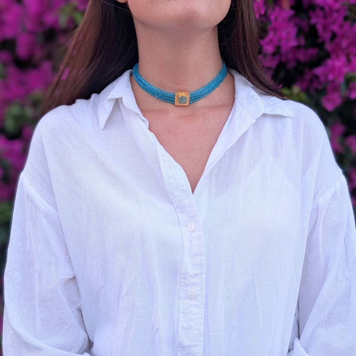 Choker de cuentas turquesa con placa dorada y piedra clara de AMANAI, con buganvilla al fondo