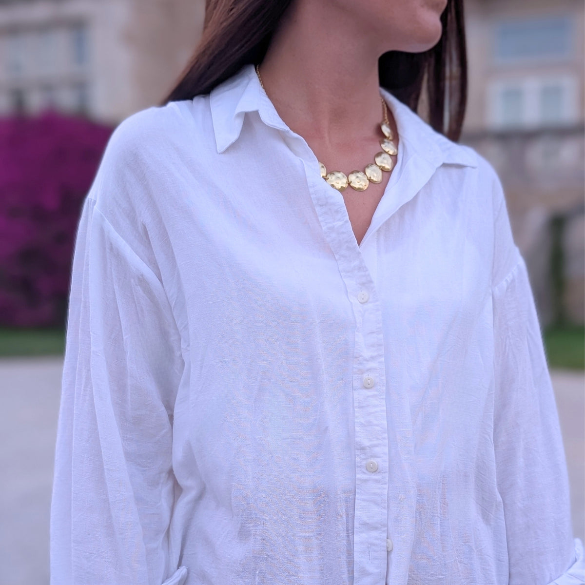 Collar de discos dorados de AMANAI sobre camisa blanca de lino, con buganvilla al fondo