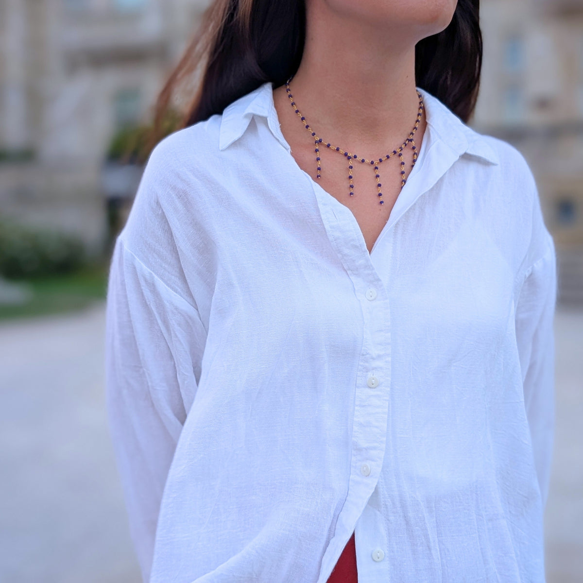 Collar dorado con flecos de piedras azules de AMANAI sobre camisa blanca en los jardines de la Magdalena