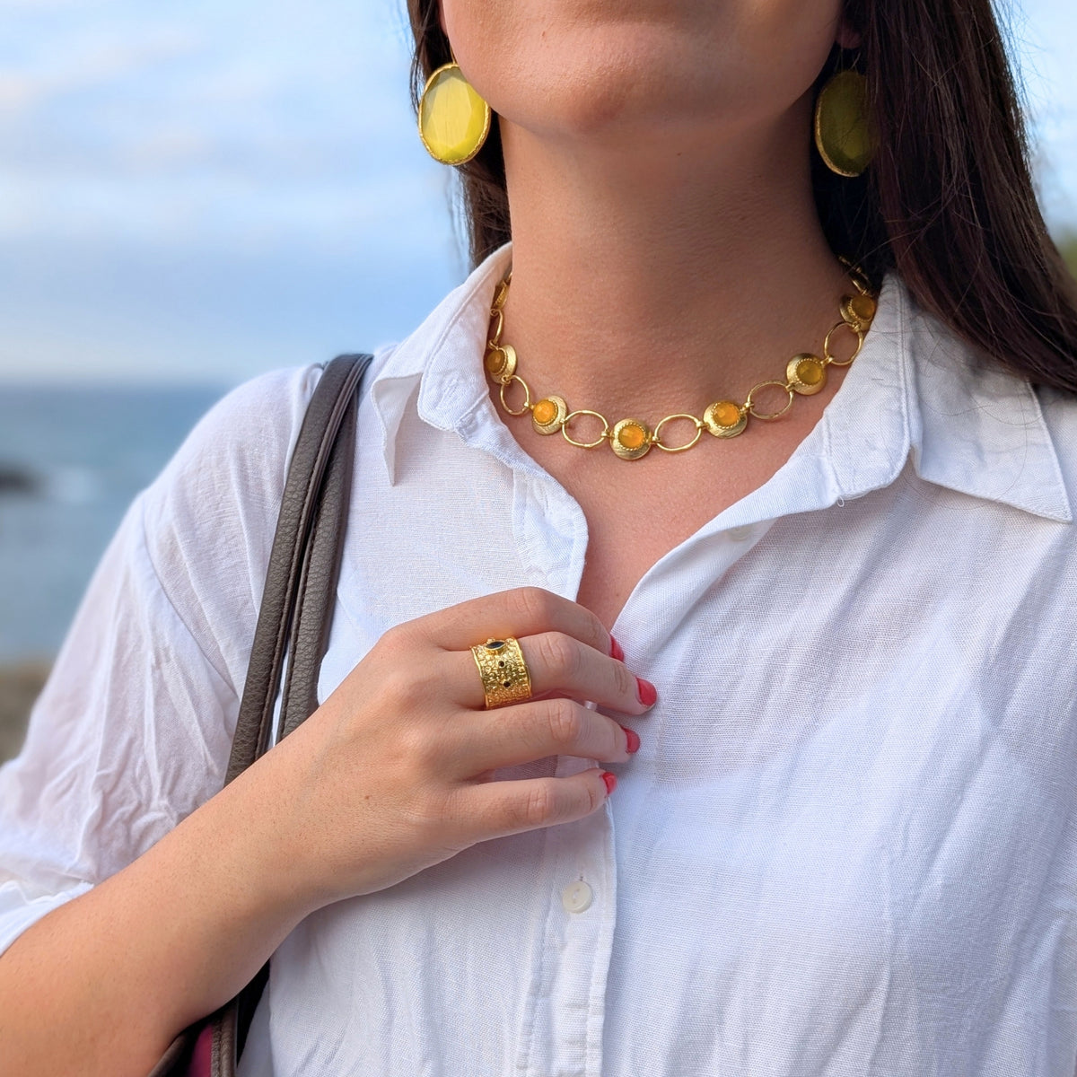Conjunto de collar dorado con piedras ámbar, pendientes ovalados y anillo ancho Neptuno de AMANAI sobre camisa blanca